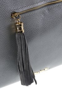 Close-up van een donkergrijze leren tas met textuur, voorzien van een gouden ritssluiting en een zwart suède franje aan de ritssluiting.