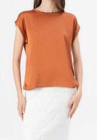 Žena v volném rustikálně zbarveném tričku s krátkým rukávem, spárovaném s bíle texturovanou sukní s vysokým pasem, stojí proti jednoduchému pozadí.