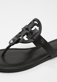 Sandale tongs noire avec un dessus texturé et découpé et des accents en métal doré. Présente une semelle lisse et mate.