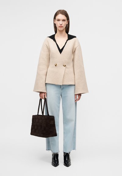 Μπεζ σακάκι από μάλλινο μείγμα με V-λαιμόκοψη και διπλό κούμπωμα, συνδυασμένο με ανοιχτό μπλε τζιν παντελόνι φαρδιάς γραμμής και σκούρα καφέ πλεκτή τσάντα ώμου.