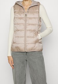 Chaleco acolchado sin mangas en beige claro, con cierre frontal de cremallera y capucha, costuras horizontales y bolsillos laterales.