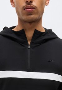 Άνδρας που φοράει μαύρη μπλούζα με κουκούλα και οριζόντια λευκή ρίγα και φερμουάρ στο μπροστινό μέρος μέχρι τη μέση, κοιτάζοντας μπροστά, με εικόνα περικομμένη από τη μύτη ως το στήθος.