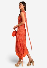 Vestido de color naranja con un estampado, con una parte trasera fluida, dobladillo en capas y un bolso naranja a juego. La modelo lleva sandalias de plataforma doradas y pulseras.