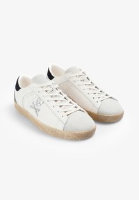 Zapatillas blancas de cuero con puntera y detalle en el talón de ante gris claro. Cuentan con diseño de cordones y suela beige texturizada. Detalle de calavera y huesos cruzados.