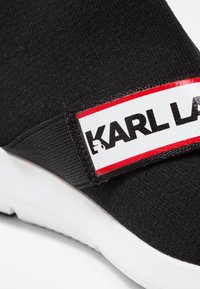 Black fabric sneaker with a white sole. Features a wide strap labeled "KARL LAGERFELD" in black text, bordered by red accents.