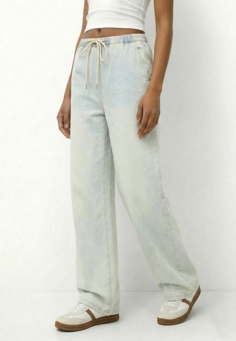 Άτομο που φοράει ανοιχτόχρωμα τζιν με φαρδιά πόδια και κορδόνι στη μέση, λευκό cropped τοπ και λευκά αθλητικά παπούτσια με καφέ σόλες.