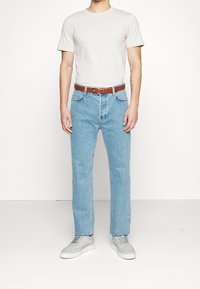 Man i en enkel ljusbeige t-shirt instoppad i ljusblå raka jeans med ett brunt bälte och ljusgrå sneakers.