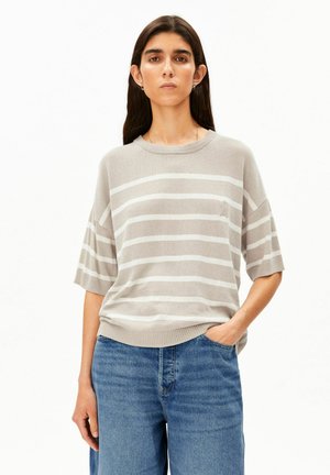 Femme aux longs cheveux foncés portant un pull à manches courtes beige et blanc rayé et un jean bleu, debout avec une main dans la poche.