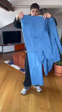 Osoba trzymająca dopasowaną jasnoniebieską marynarkę i spodnie w salonie z drewnianą podłogą i pomarańczowym umeblowaniem.