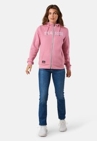 Helle rosa Zip-Up-Hoodie mit "SEASIDE" in Weiß bedruckt, mit vorderen Taschen und Kordelzugkapuze, kombiniert mit blauen Jeans und weißen Sneakern.