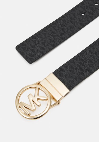 Ceinture noire texturée avec une boucle circulaire dorée brillante portant les initiales "MK". La sangle présente un motif subtil et plusieurs trous d'ajustement.