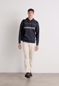 Hanorac cu glugă de culoare bleumarin închis, cu un logo tonal, asortat cu pantaloni off-white și pantofi negri, purtat cu o geantă de umăr. Design simplu.