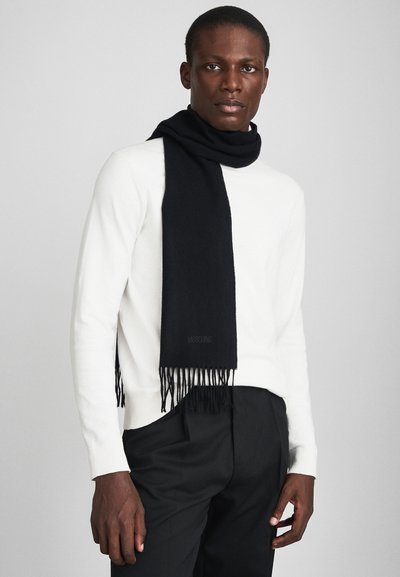 Melnā šalle ar frizētām malām, izgatavota no mīkstas auduma, uzvilkta virs baltā džempera. Zīmola logo ir diskreti attēlots uz šalles.