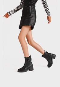 Stivali neri alla caviglia con un tacco spesso, in materiale di pelle, con un pannello laterale texturizzato e una fascia decorativa con dettaglio a fibbia.