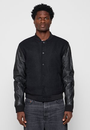 Mann trägt eine schwarze College-Jacke mit Wollkörper und Ledärmeln, steht vor einem einfarbigen grauen Hintergrund und blickt nach vorn.