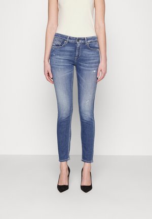 Kobieta w wyblakłych, obcisłych niebieskich dżinsach z drobnymi przetarciami i czarnych szpilkach z zaokrąglonym noskiem, stojąca na jednolitym tle.