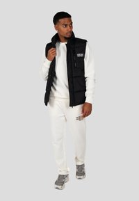 Gilet imbottito nero con chiusura con zip e logo, indossato sopra un maglione bianco e pantaloni abbinati. Scarpe da ginnastica grigie completano l'outfit.