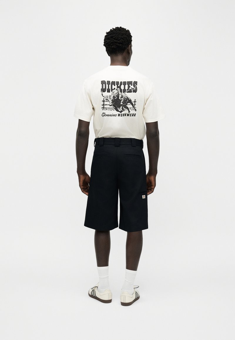 Uomo che indossa una t-shirt bianca con grafica di un toro, pantaloncini neri fino al ginocchio, calzini bianchi e scarpe da ginnastica beige, in piedi di spalle.