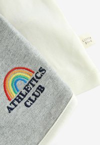 Grijze katoenen stof met een geborduurde meerkleurige regenboog en marineblauwe tekst "ATHLETICS CLUB." De andere kant is crèmekleurig met een label.