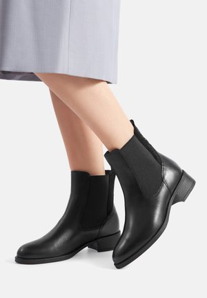 Czarne skórzane botki za kostkę z elastycznymi panelami po bokach, niskim obcasem, gładkim wykończeniem i zaokrąglonym noskiem. Noszone z jasnoszarą spódnicą.