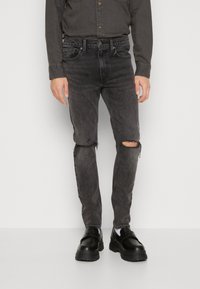 Jeans skinny negre, deteriorate, cu detalii de genunchi destramate, realizate din țesătură de denim, asortate cu pantofi masivi negri și o cămașă gri cu nasturi.