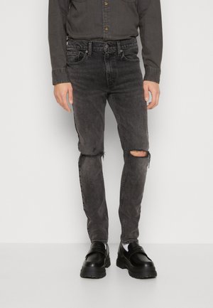 Svarte slitte skinny jeans med frynsete knedetalj, laget av denimstoff, kombinert med kraftige svarte sko og en grå skjorte med knapper.