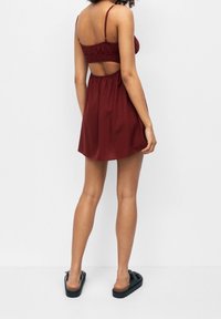 Vínové šaty bez rukávů se staženým pasem a otevřenými zády, vyrobené z hladké látky, doplněné černými nazouváky.