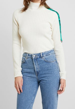 Maglione bianco a coste con collo alto realizzato in tessuto morbido, con una fascia turchese con testo nero lungo la spalla, abbinato a jeans azzurri chiari.
