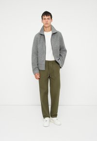 Szary wełniany płaszcz z wysokim kołnierzem, zestawiony z oliwkowymi spodniami i białymi sneakersami. Prosty design, bez widocznych wzorów czy akcentów.