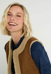 Mujer sonriente con cabello corto rubio, que lleva pendientes de aro de oro, una camisa azul marino bordada con flores y un chaleco de piel de oveja marrón.