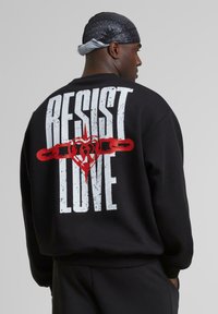 Black sweatshirt featuring large white text "RESIST" and "LOVE" with a red heart and chain graphic on the back; crew neck and relaxed fit.