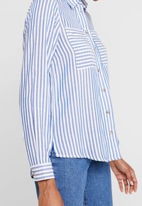 Chemise boutonnée à rayures verticales bleu et blanc, avec une poche poitrine, des manches longues et un ourlet arrondi. Confectionnée dans un tissu léger.