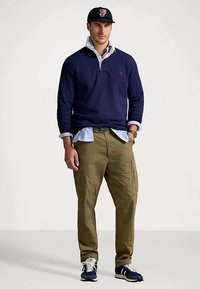 Άντρας που φορά ναυτικά μπλε μπλουζάκι ράγκμπι πάνω από ανοιχτό μπλε πουκάμισο με γιακά, ελαιόχρωμο cargo παντελόνι, ναυτικά αθλητικά παπούτσια και μαύρο καπέλο μπέιζμπολ, στέκεται μπροστά σε άσπρο φόντο.