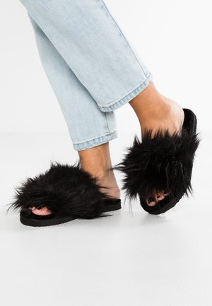 Sandales mules noires en fourrure avec une texture douce, design à bout ouvert, et semelle en caoutchouc, portées avec un jean bleu clair retroussé.