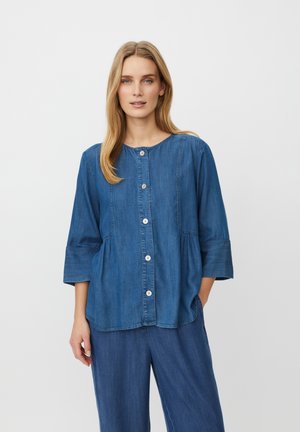 Žena s dlouhými blond vlasy, oblečená v zapnuté džínové košili s tříčtvrtečními rukávy a odpovídajících širokých džínových kalhotách, stojí před bílým pozadím.