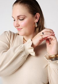 Beige trui met gerimpelde mouwen, gedragen met gelaagde gouden sieraden en ringen. Glad oppervlak, met een aansluitend ontwerp dat de vorm flatteert.