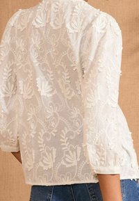 Blusa bianca ricamata con motivi floreali, maniche a sbuffo corte e un tessuto trasparente testurizzato. Vestibilità casual con lunghezza leggermente accorciata.