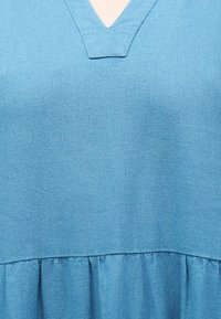 Ανοιχτό μπλε ρούχο με V-λαιμόκοψη και λεπτομέρεια ρυμουλκούμενου υφάσματος κάτω από το στήθος, που δείχνει την υφή και το ράψιμο στο πλεκτό υλικό.