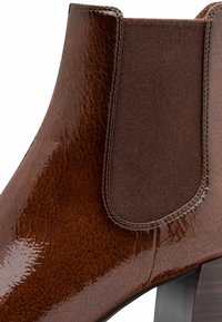Stivale alla caviglia in pelle marrone con finitura lucida, pannello laterale elasticizzato e tacco chunky. Presenta una superficie testurizzata e dettagli in materiali contrastanti.