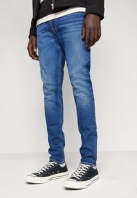 Jeans de denim azul con un corte ajustado, que presentan un ligero desvanecimiento, costuras en contraste y bolsillos frontales; combinados con zapatillas negras y cordones blancos.