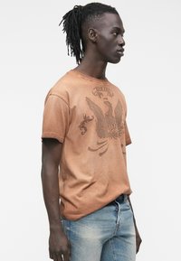 Camiseta gráfica marrón con un diseño texturizado de un águila y la palabra "DIESEL." Combínala con jeans azules desgastados, con corte recto en la pierna.