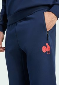 Navy jogger pants in a smooth fabric with a zippered pocket, featuring a bright red rooster logo on the side.