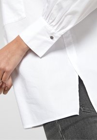 Weißes Hemd mit langen, gepufften Ärmeln und abgerundetem Saum. Verfügt über einen kleinen, dekorativen silbernen Knopf am Bündchen.