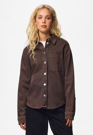 Jeune femme aux longs cheveux blonds portant une chemise marron à boutons avec une poche poitrine et un pantalon noir sur fond blanc.