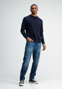 Navy gebreide trui met ribbelmanchetten, gecombineerd met lichtblauwe spijkerbroek en zwarte sneakers, met witte rubberen zolen en zwarte veters.