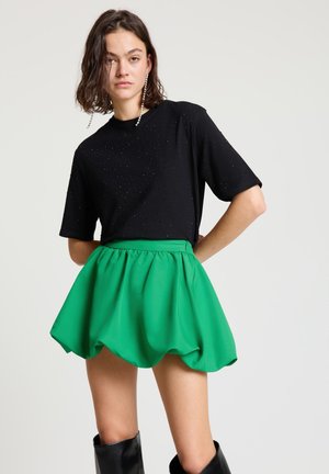 Donna che indossa una maglietta a maniche corte nera con dettagli di perline, una mini gonna verde brillante arricciata, lunghi orecchini di perle e stivali neri al ginocchio.