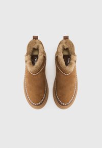 Un paio di scarpe in suede marrone da infilare per l'inverno, con spessa fodera in pelliccia beige e cuciture decorative bianche sul bordo della suola.