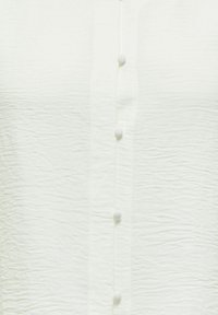 Tissu blanc froissé avec une rangée verticale de petits boutons recouverts le long d’une patte de boutonnage centrale.