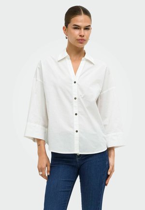 Femme aux cheveux foncés attachés, portant une chemise blanche ample à boutons avec de larges manches et un jean bleu sur un fond uni.