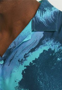 Primer plano de una persona que lleva una camisa de patrón azul y turquesa con cuello y detalle de botones, mostrando el área del cuello y los hombros.